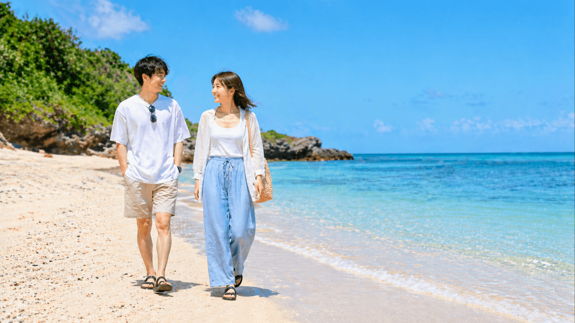 沖縄の透き通る青い海と白い砂浜を、笑顔で散歩する若いカップルの様子。