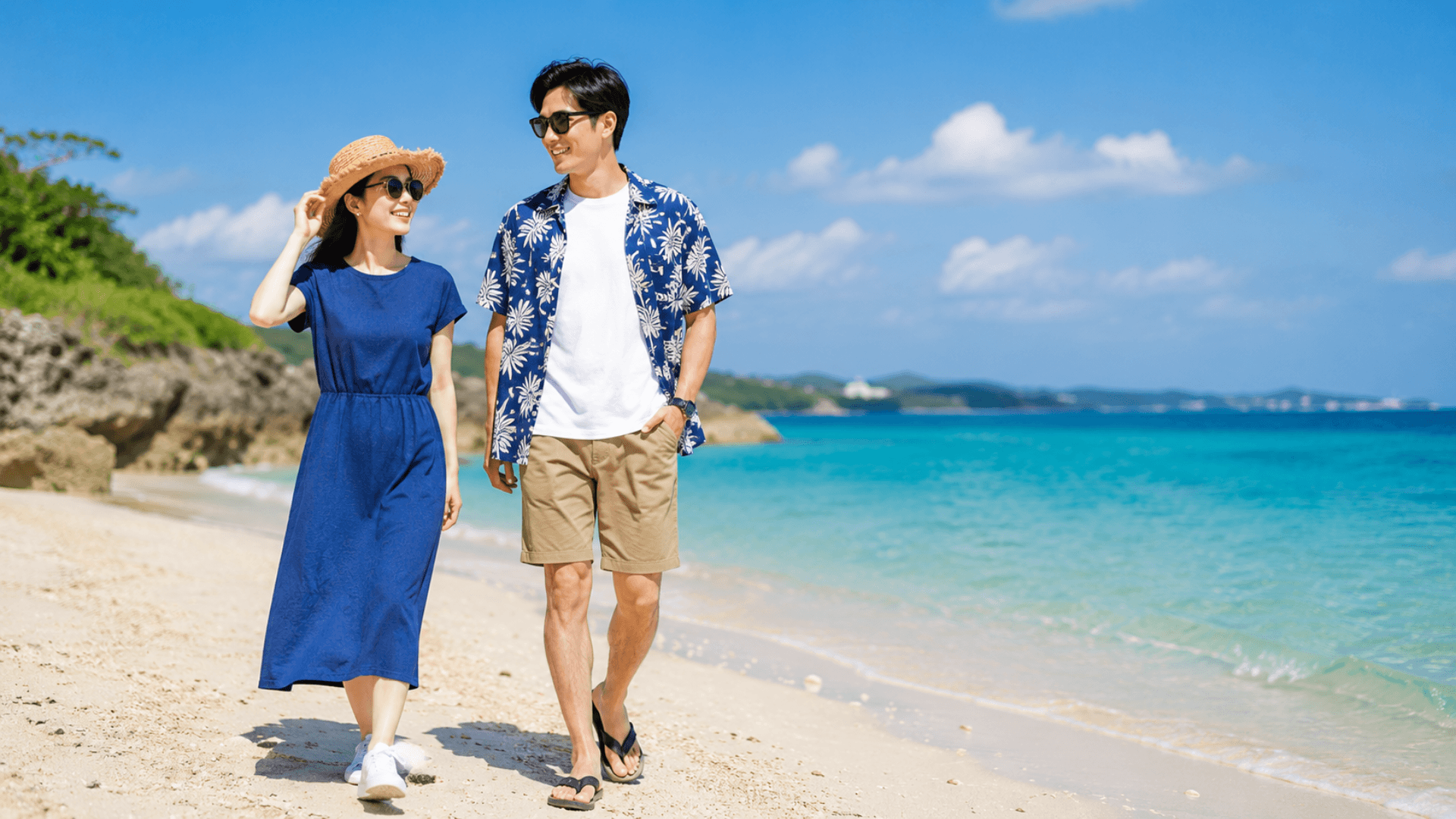 6月の沖縄の海辺を歩くカップルのイメージ。男性は青い沖縄風の柄シャツに白Tシャツ、女性は涼しげな半袖ワンピースに麦わら帽子、サングラス、白いスニーカーを着用。